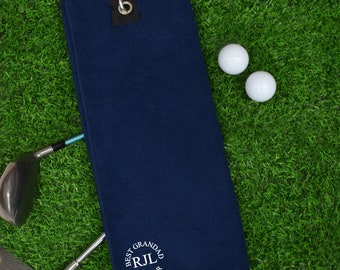 May include: A dark blue golf towel with a white embroidered circle that says "Best Grandad RJL By Par". The towel has a loop for hanging and is lying on a green surface with two white golf balls.
