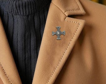 Puede incluir: Un broche plateado en forma de cruz con un acento central similar a una perla está prendido a una blazer color beige. La blazer se lleva sobre un suéter azul oscuro. El broche es un accesorio clásico.