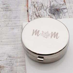 Könnte beinhalten: Eine silberfarbene, runde Schmuckdose mit dem Wort "MOM" in hellvioletter Schrift, mit einem Blumendesign zwischen den Buchstaben. Die Dose ist geschlossen und hat einen kleinen Verschluss. Der Hintergrund ist eine helle Holzoberfläche.
