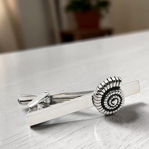 May include: A silver-toned metal tie clip featuring a smooth, rectangular bar design accented with a detailed, raised ammonite fossil motif on one end. The accessory has a polished, reflective finish and a spring-loaded mechanism for secure attachment.