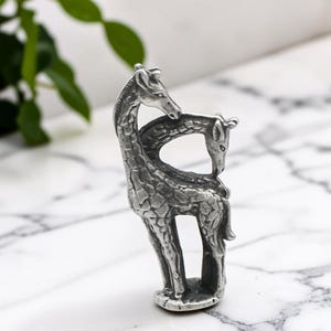 May include: A silver-toned metal figurine depicting two giraffes standing together. The taller giraffe stands upright while the smaller giraffe is positioned beside it with its head lowered, both featuring detailed textured patterns on their bodies.