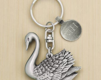 May include: A silver-colored swan keychain with a round tag engraved with the initials "MH". The swan has detailed feathering and is attached to a key ring with a swivel clasp. The background is a light wood grain.