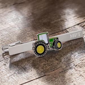 Puede incluir: Pinza de corbata plateada con un diseño de tractor verde y amarillo. El tractor tiene neumáticos negros y una cabina blanca. Las iniciales "MPT" están grabadas en la pinza. La pinza está sobre una superficie de madera.