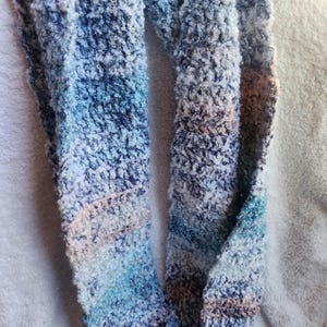 May include: A soft, textured infinity scarf in shades of blue, white, and a hint of peach. The scarf is made of a plush, looped yarn, creating a cozy and warm accessory. The color palette evokes a serene, wintery feel.