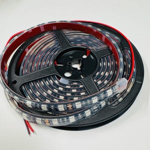Puede incluir: Un rollo de tira de luz LED negra con respaldo adhesivo 3M. La tira est&aacute; enrollada alrededor de un carrete de pl&aacute;stico negro con un cable rojo y negro que sale del extremo.