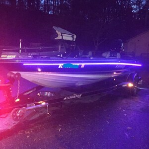 Puede incluir: Un barco de pesca blanco y negro con una franja de luz azul y morada a lo largo del lateral. El barco est&aacute; en un remolque con el nombre "STRATOS" impreso en el lateral. El barco tiene un dise&ntilde;o blanco y negro con un portaca&ntilde;as de pesca blanco y negro en la parte delantera.
