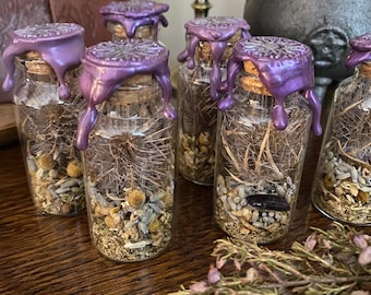 May include: Several small glass bottles filled with dried herbs and flowers, sealed with corks and dripping purple wax. The bottles are arranged on a wooden surface, with additional dried herbs in the foreground. A cauldron and chalice are in the background.