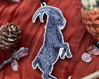 Könnte beinhalten: Ein Holzanhänger mit einer detaillierten schwarzen Tuschezeichnung einer Ziege, die auf ihren Hinterbeinen steht. Die Ziege hat gebogene Hörner und strukturiertes Fell. Der Text paintedwitchdesigns ist vertikal an der Seite aufgedruckt.