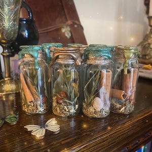 May include: A collection of small glass bottles with cork stoppers and a green wax seal. Each bottle contains dried herbs, flowers, and crystals. The bottles are arranged in a row on a wooden surface.
