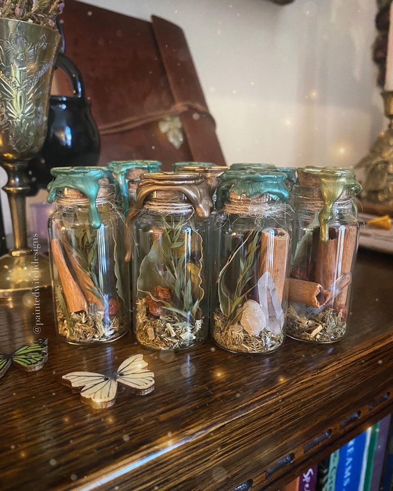 May include: A collection of small glass bottles with cork stoppers and a green wax seal. Each bottle contains dried herbs, flowers, and crystals. The bottles are arranged in a row on a wooden surface.