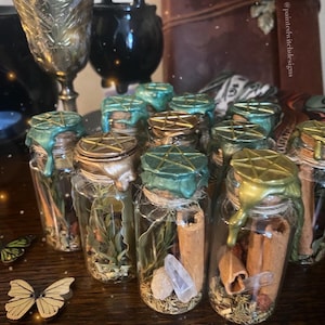 May include: A collection of small glass bottles with cork stoppers sealed with green and gold wax. Each bottle contains various herbs, crystals, and other natural ingredients. The bottles are arranged in a row on a wooden surface. @paintedwitchdesigns