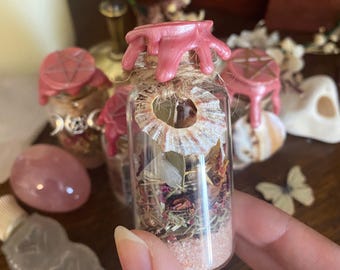 May include: A small glass bottle filled with pink sand, dried herbs, and crystals. The bottle is sealed with a pink wax top and a seashell around the neck. Other bottles and crystals are in the background.