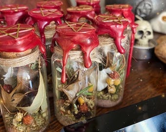 May include: A collection of small glass jars filled with dried herbs and botanicals. Each jar is sealed with a red wax top featuring a gold pentagram. The jars are tied with twine and sit on a wooden surface.