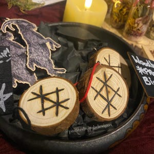 May include: Circular wooden ornaments with a natural, rustic appearance. Each ornament has a black symbol burned into the wood. The ornaments are displayed in a dark metal tray with black paper. Some have a red string attached.