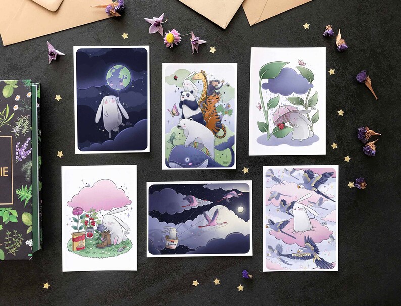 May include: Set of six illustrated cards featuring a white rabbit in different scenes. The cards are printed on white cardstock and have a matte finish. The scenes include a rabbit flying in the night sky, a rabbit under a cloud, a rabbit in a forest, a rabbit in a field, a rabbit in a garden, and a rabbit in a sky with birds.