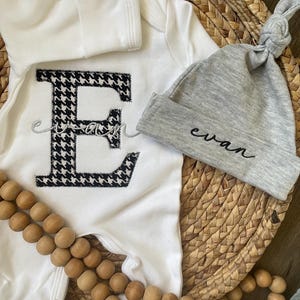 May include: A white baby onesie with a large black and white houndstooth letter "E" and the name "Evan" embroidered on a gray knotted hat. A wooden bead garland is draped across the image, which is set on a woven basket.