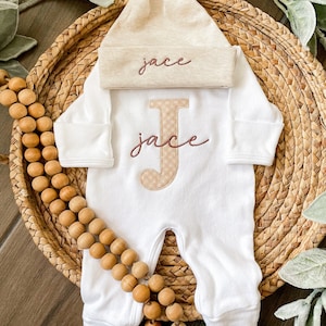 May include: A white baby onesie and matching knotted hat set. The onesie features a large, embroidered letter "J" with a tan and white checkered pattern, and the name "Jace" embroidered in a cursive font. The hat also has the name "Jace" embroidered on it.