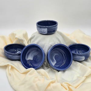 May include: A set of five deep blue ceramic bowls with a smooth, glossy finish. Each bowl features a distinct white brushstroke pattern along the exterior or interior, showcasing a handcrafted aesthetic with visible circular textures inside.