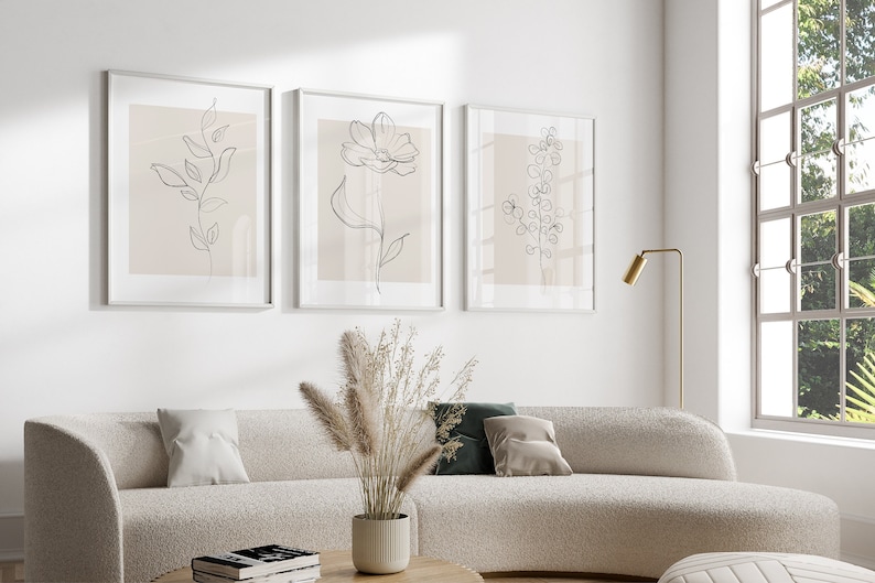 May include: Three framed minimalist line drawings of flowers and leaves on a white wall. The drawings are in black and white and have a light pink background. The frames are white and the drawings are arranged in a row.