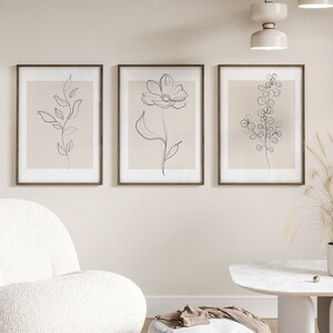 May include: Three framed minimalist line drawings of flowers and plants on a beige wall. The drawings are in black and white and are set against a light beige background. The frames are brown wood.