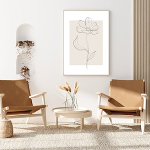 May include: A minimalist line drawing of a flower in a light brown frame hangs on a white wall above a light brown rug. Two brown leather chairs are placed on either side of a small wooden table with a vase of dried grass.