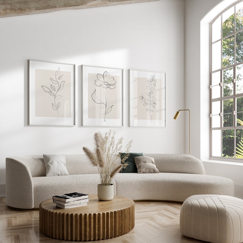 May include: Three minimalist line art prints of floral designs in black and white on a light pink background, framed in white and hung on a white wall above a white curved sofa with a wooden coffee table and a fluffy white ottoman.