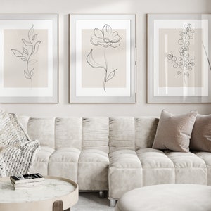 May include: Three framed minimalist line drawings of plants on a light beige wall above a white tufted sofa with two pillows. The sofa is in a living room setting with a coffee table and a throw blanket.