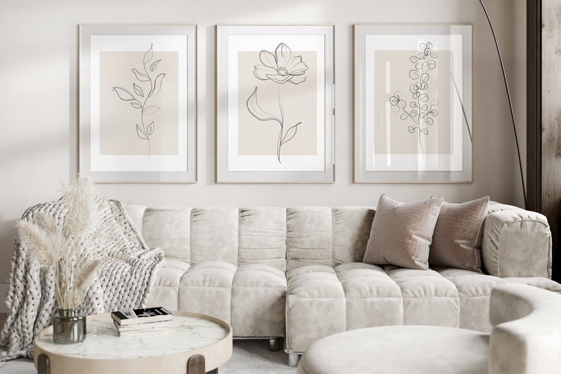May include: Three framed minimalist line drawings of plants on a light beige wall above a white tufted sofa with two pillows. The sofa is in a living room setting with a coffee table and a throw blanket.