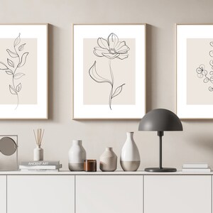 May include: Three minimalist line art prints of floral designs in black and white on a beige background. The prints are framed in a light wood frame and are displayed on a white shelf with decorative vases and a mirror.