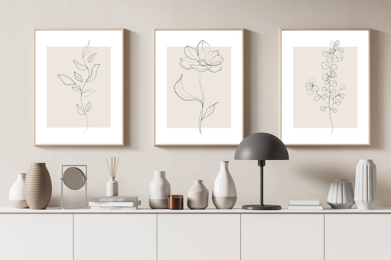 May include: Three minimalist line art prints of floral designs in black and white on a beige background. The prints are framed in a light wood frame and are displayed on a white shelf with decorative vases and a mirror.