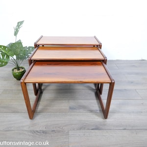 May include: A set of three nested tables made of polished brown wood with a smooth, rectangular surface and rounded edges on the legs. The tables are stacked together, showing a consistent grain pattern and a warm, medium-brown finish throughout.