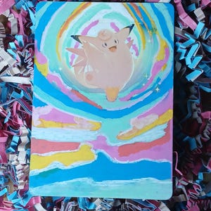 May include: A rectangular card featuring a hand-painted illustration of a light pink, round creature with pointed ears and a small tail. The creature is centered within a swirling, colorful aura of blue, yellow, and pink paint strokes.