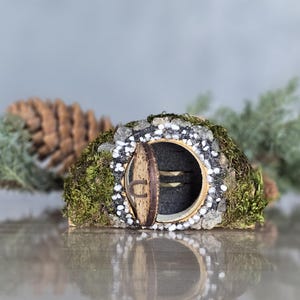 Op de afbeelding: Een miniatuur fee&euml;nhuis met een ronde deur en een mossige buitenkant. Het huis is versierd met witte stenen en is omgeven door groen gebladerte.
