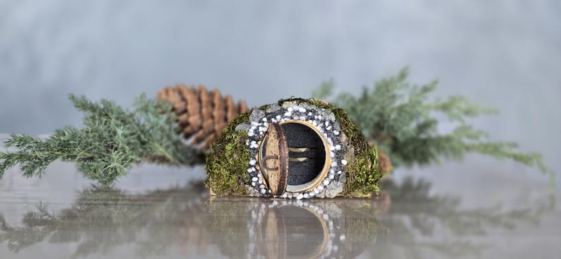 Op de afbeelding: Een miniatuur fee&euml;nhuis met een ronde deur en een mossige buitenkant. Het huis is versierd met witte stenen en is omgeven door groen gebladerte.