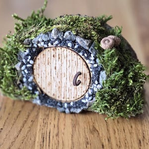 Op de afbeelding: Een miniatuur hobbit-hol met een houten deur, omringd door mos en kleine stenen. De deur heeft het nummer "6" erop.