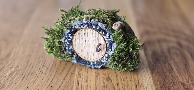 Op de afbeelding: Een miniatuur hobbit-hol met een houten deur, omringd door mos en kleine stenen. De deur heeft het nummer "6" erop.