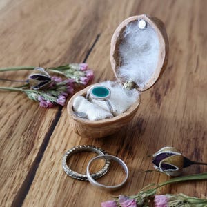 Puede incluir: Una caja de anillo hecha de una cáscara de nuez partida por la mitad con un forro de tela blanca. Un anillo verde está dentro de la caja. Dos bandas plateadas están sobre una superficie de madera con flores secas.