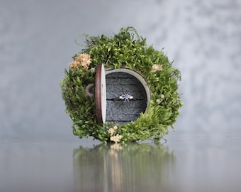 May include: A circular ring box covered in vibrant green moss with small light-colored dried flowers. The box features a round wooden door with a small handle, opening to reveal a dark gray interior slot holding a silver ring with a blue stone.
