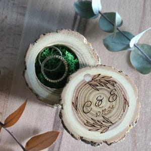May include: A wooden ring box with a natural bark finish. The lid is engraved with the initials "C & E" and the date "10.19.24". The box is filled with green moss and two gold rings.