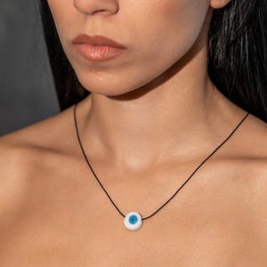 May include: A minimalist necklace with an eye-shaped pendant in white and blue, strung on a slender black cord. The pendant's design is a white base with a blue centre. This necklace is a subtle yet stylish piece of jewellery.
