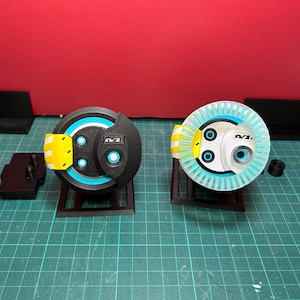 May include: Two black and white toy robots with blue and yellow accents. One robot has a circular design with a blue ring and the other has a white circular design with a blue ring. Both robots are on black stands.