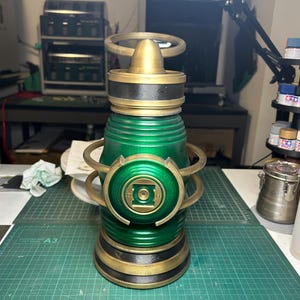 May include: A metallic green and gold lantern prop featuring a central circular emblem with a square cutout. The cylindrical object has a ribbed body, a top handle ring, and a wide base, finished with dark gold accents and a glossy green surface.