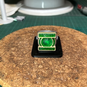 Könnte beinhalten: Ein Green Lantern Ring mit einem metallisch silbernen Band und einer rechteckigen Vorderseite, die ein erhabenes, glitzerndes grünes kreisförmiges Emblem in einem goldfarbenen Rahmen zeigt, montiert auf einem kleinen schwarzen quadratischen Ständer.