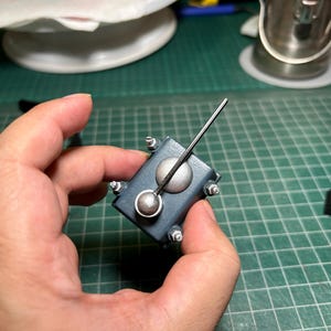 May include: A small, dark blue rectangular object with a silver-coloured dial and a long, thin, black rod extending upwards. The object has silver-coloured bolts on each corner. The object is held in a hand.