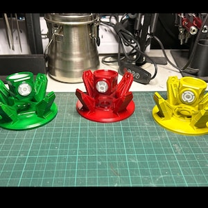 Op de afbeelding: Drie 3D-geprinte Green Lantern-ringen, elk met een andere gekleurde basis: groen, rood en geel. De ringen zijn ontworpen om een Green Lantern-ring te houden.