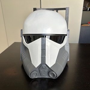 Puede incluir: Un casco blanco y gris con visera, probablemente para cosplay o disfraz. El casco tiene una parte superior redondeada y elementos angulares, con una franja central gris y secciones inferiores grises. El casco está sobre una superficie oscura.