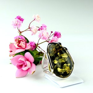 May include: A silver ring with a large, oval-shaped green amber stone. The stone has a unique, marbled pattern and is set in a simple, elegant band. The ring is surrounded by pink flowers.