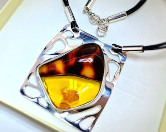 May include: A silver pendant necklace with a rectangular frame and an amber stone. The amber is a mix of yellow and brown hues. The necklace has a black cord and a silver clasp.