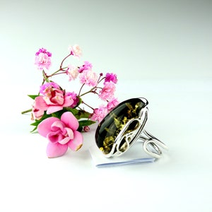 May include: A silver ring with a large green amber stone set in a delicate, swirling design. The ring is displayed on a white surface with pink flowers.