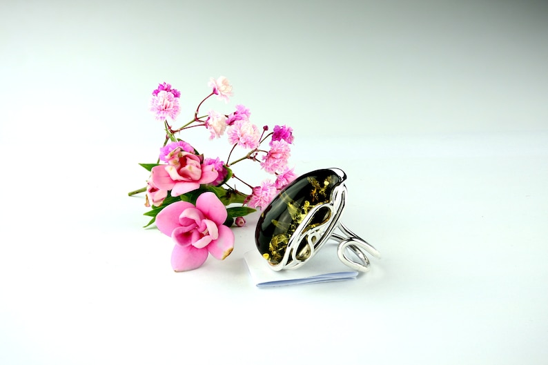 May include: A silver ring with a large green amber stone set in a delicate, swirling design. The ring is displayed on a white surface with pink flowers.